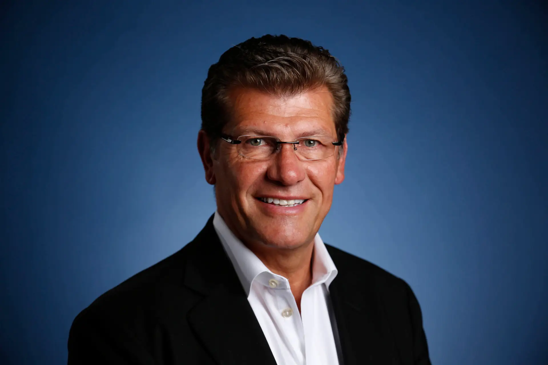 Smiling man in glasses wearing a dark jacket and white shirt against a blue background.