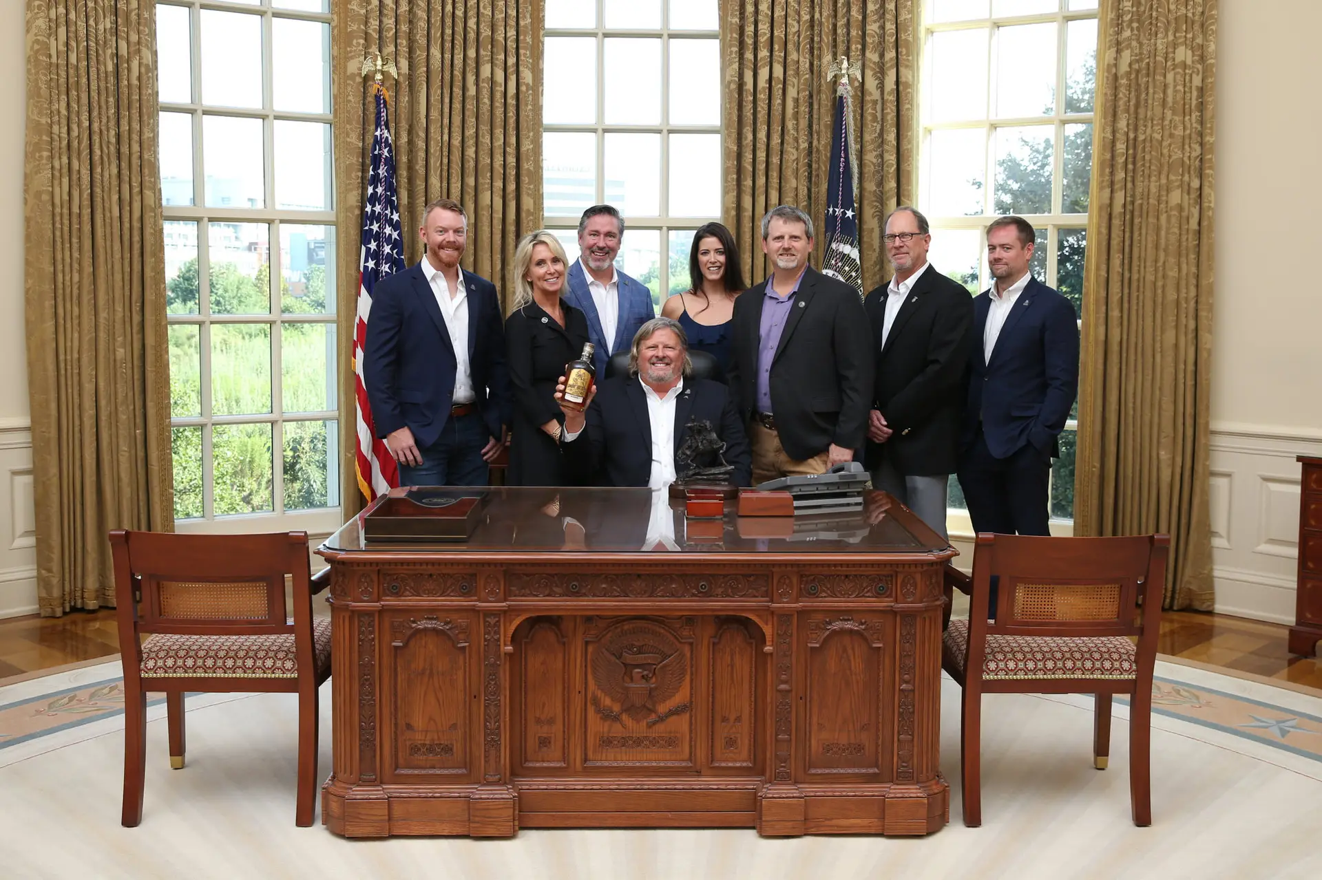 Group posing around an ornate wooden desk in a room with tall windows and flags.