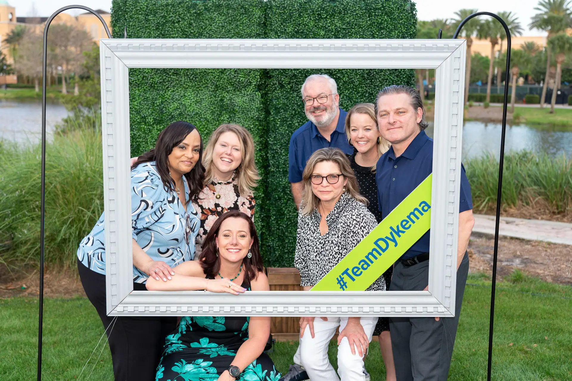 Seven people posing outdoors inside a large decorative frame with a Team Dykema banner.