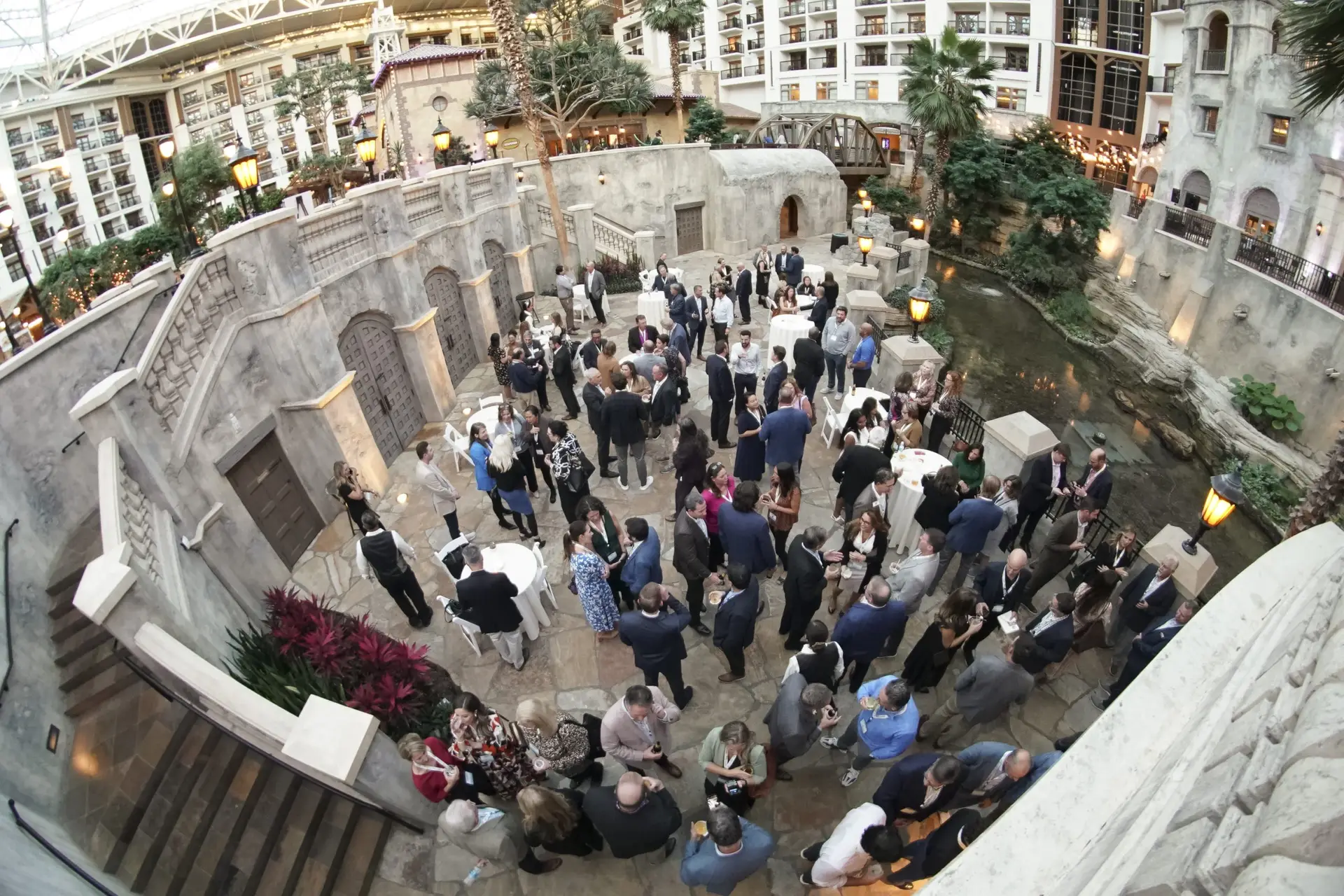 Overhead view of people gathering around cocktail tables in a planted indoor atrium.