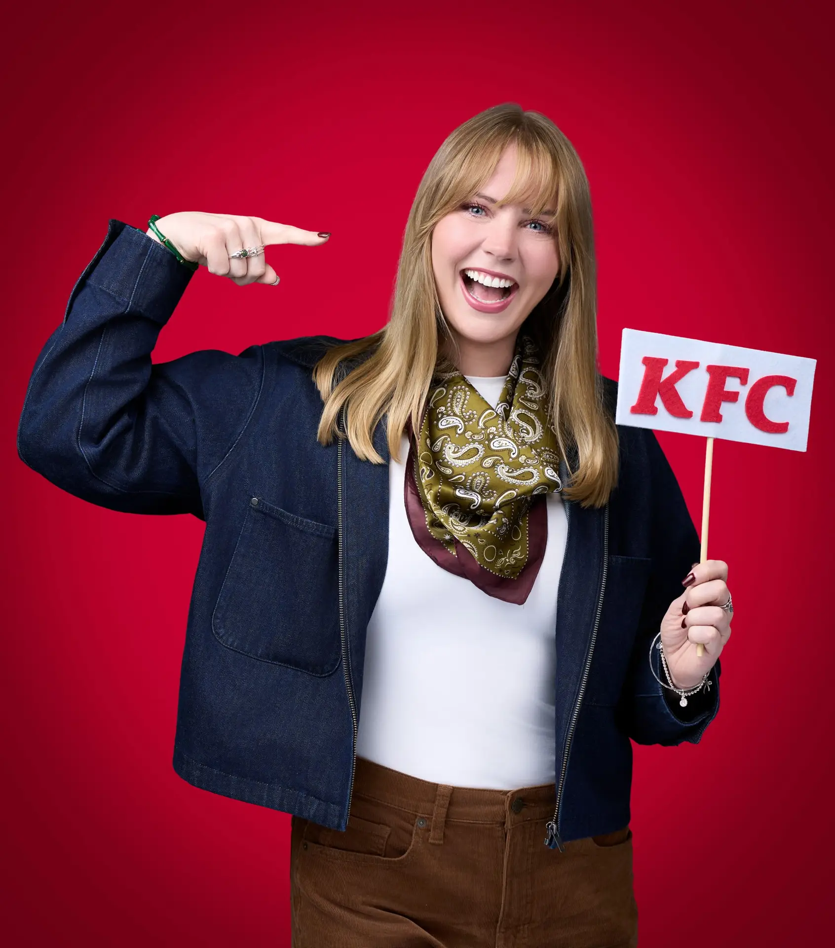 Woman in a navy blazer and patterned scarf holding a KFC sign and gesturing toward herself.