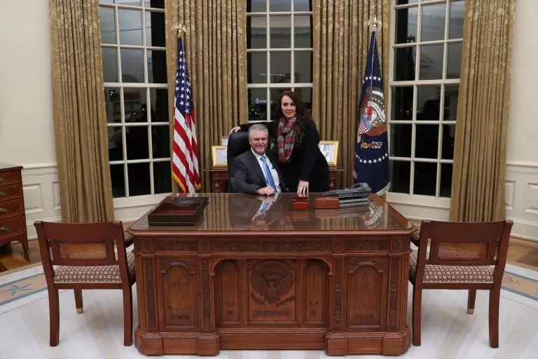 Man seated at an ornate wooden desk with a woman standing beside him between two flags.