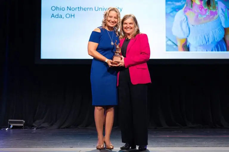Two women in blue and pink outfits holding an award together onstage.