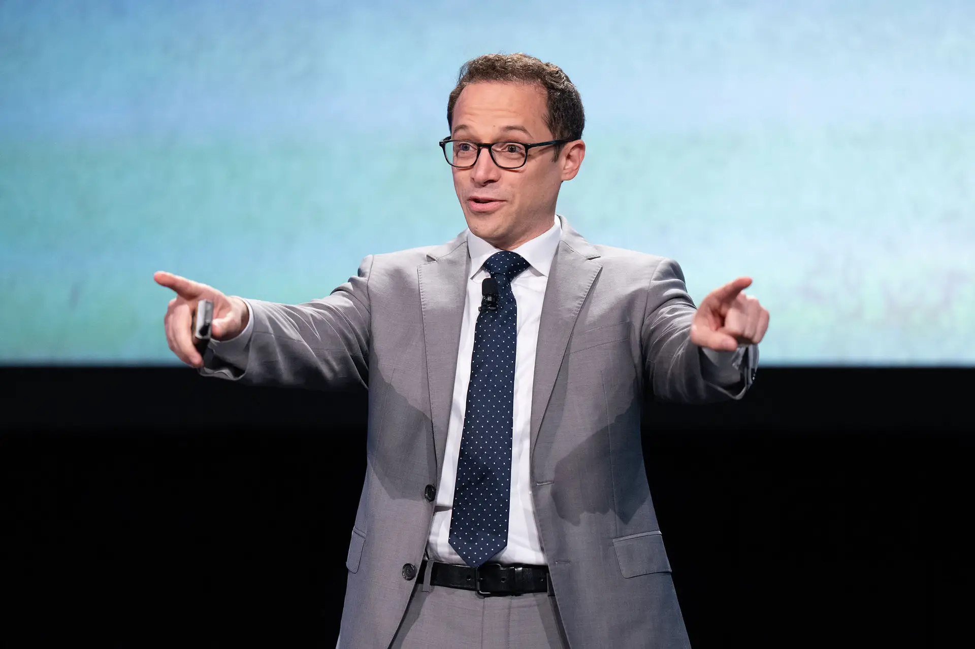 Man in glasses and a gray suit pointing with both hands during an onstage presentation.