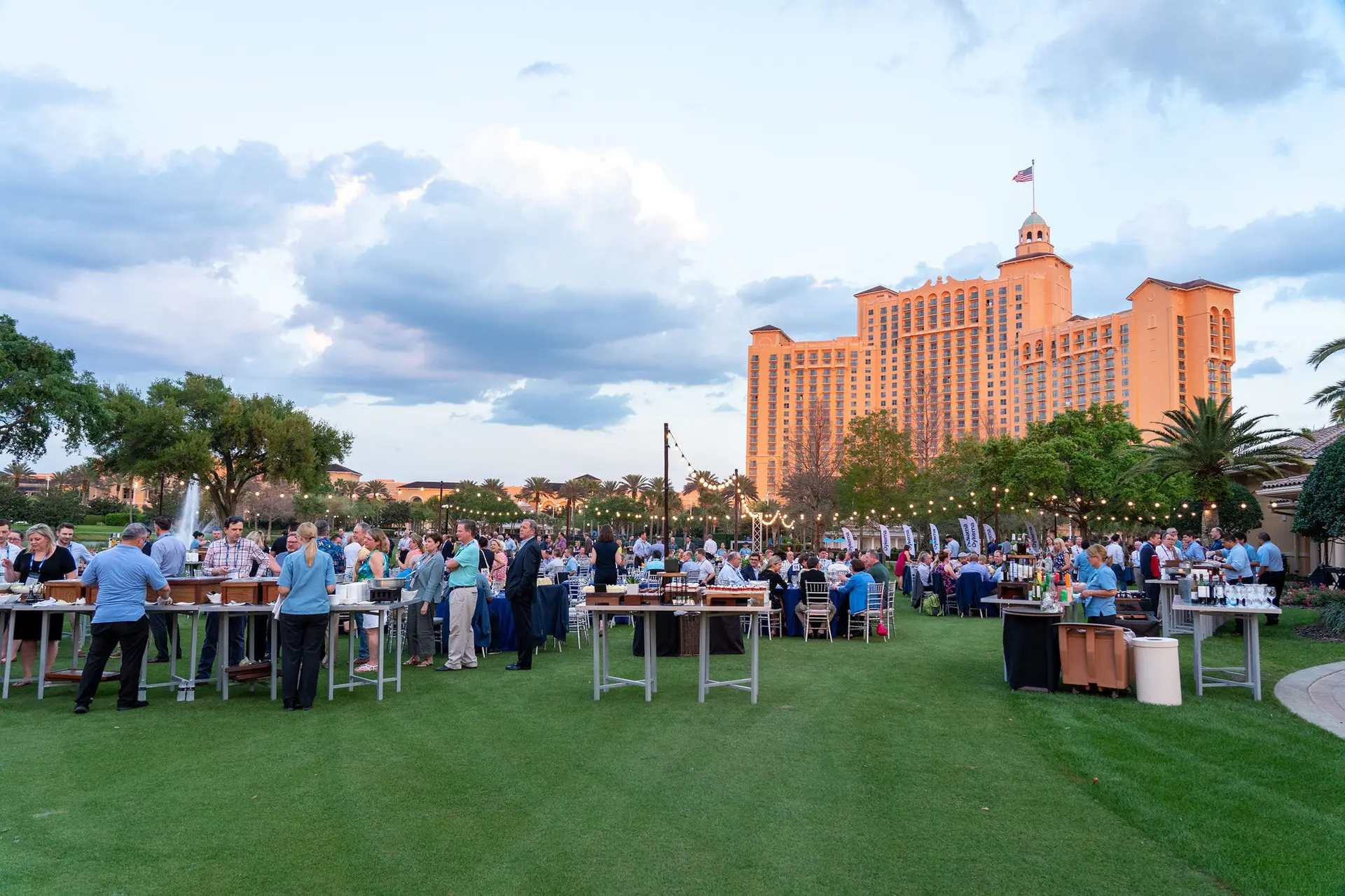 Guests gathering around cocktail tables on a lawn with a large building in the background.