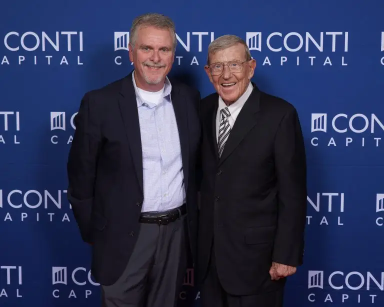 Two men in suits posing together in front of a blue Conti Capital backdrop.