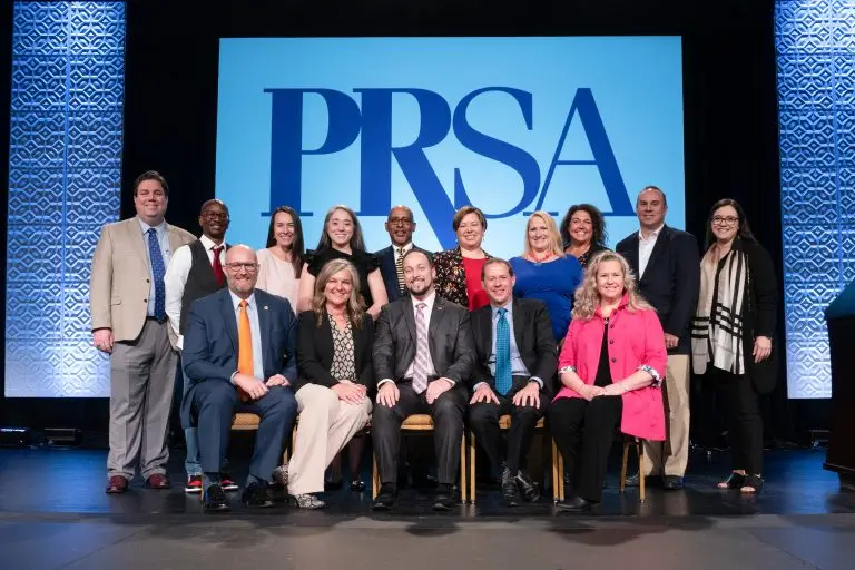 Group of people posing in two rows onstage beneath a large PRSA logo.