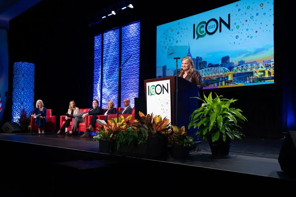 Speaker at a lectern on a blue-lit stage with panelists seated nearby and ICON signage behind.