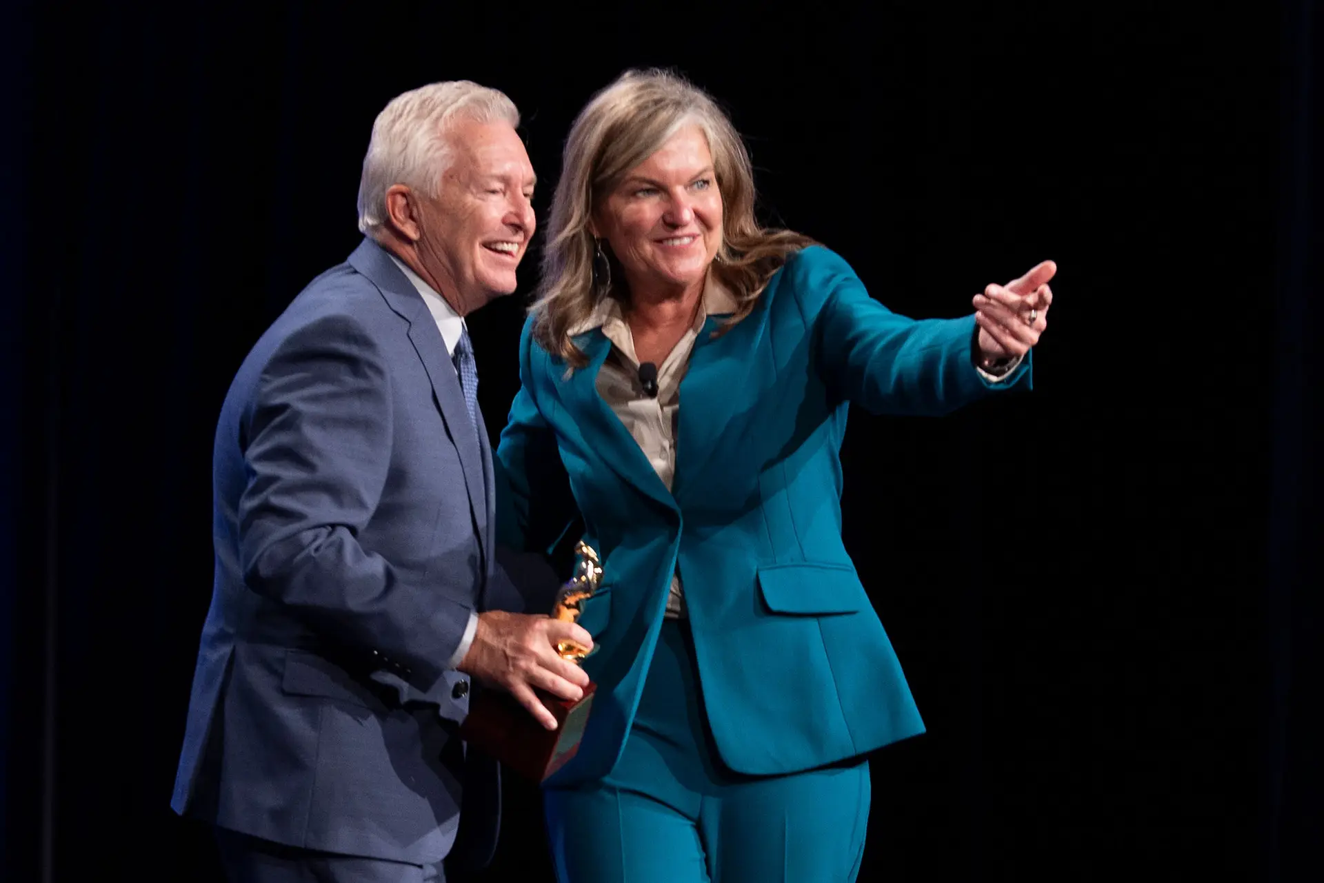 Woman in a teal suit gesturing beside a man in a blue suit holding an award.