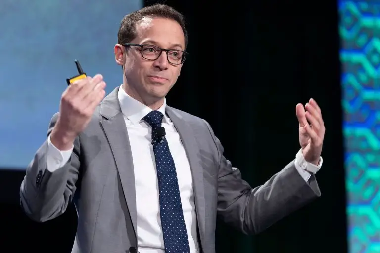 Man in glasses and a gray suit gesturing with both hands during a presentation.