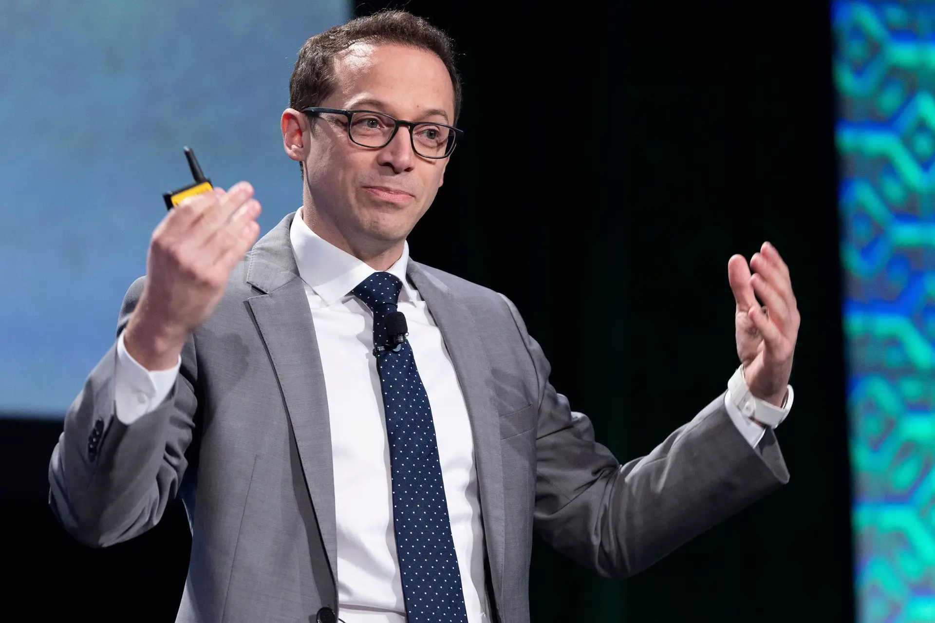 Man in glasses and a gray suit gesturing with both hands during a presentation.