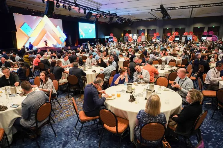 Large crowd seated at round tables in a ballroom facing a brightly lit stage.