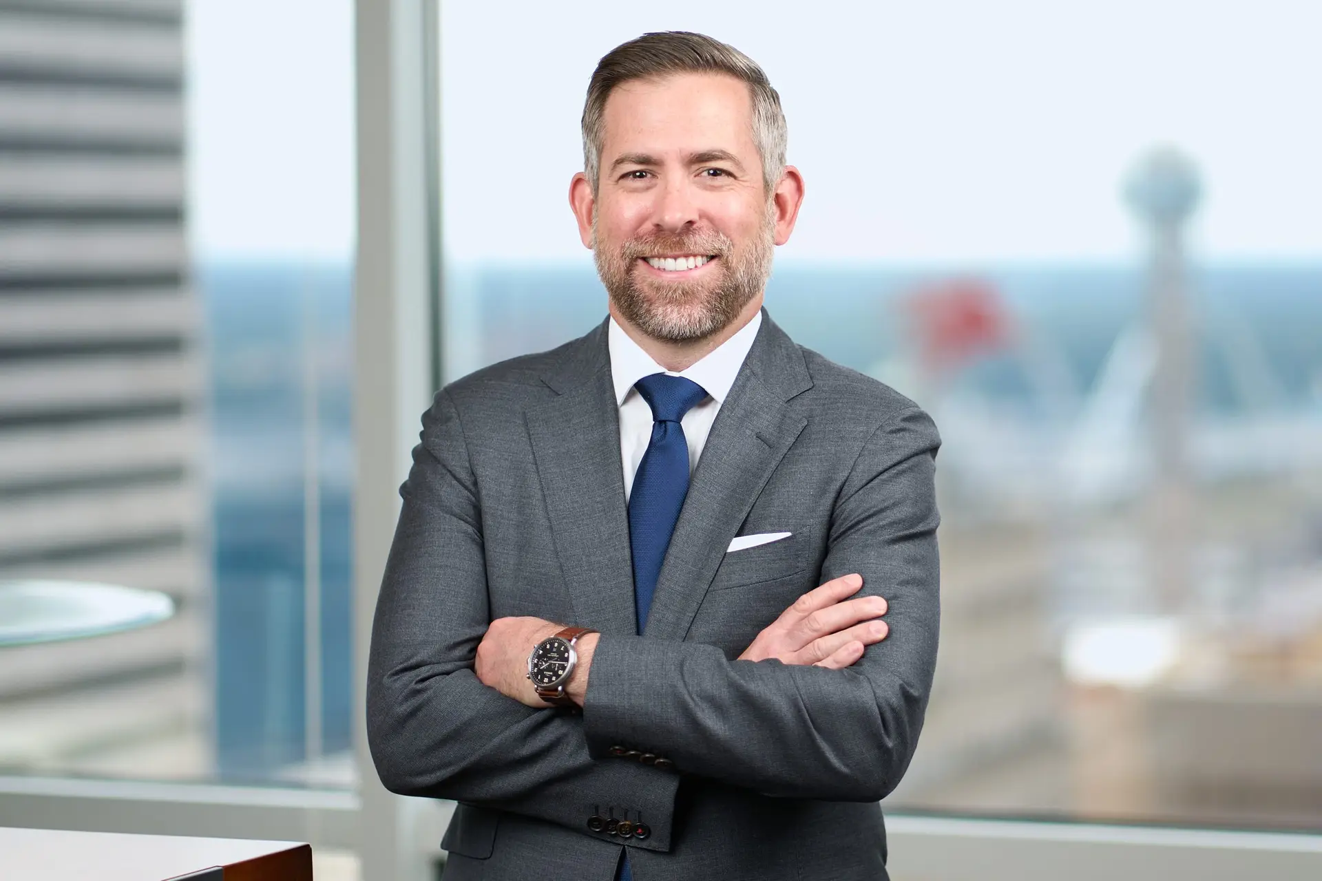 Smiling man in a gray suit and blue tie standing with arms crossed beside office windows.