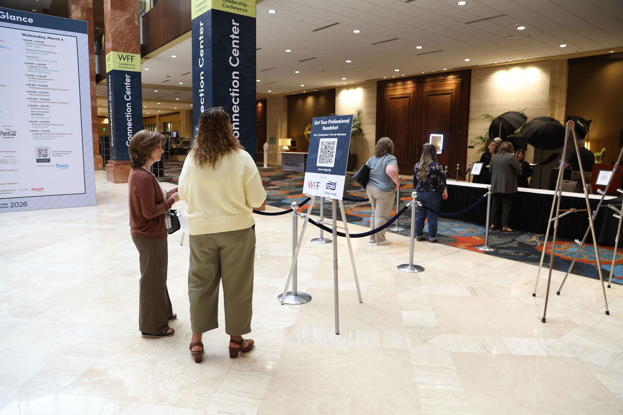 Attendees near a sign-up sign for professional headshots at the WFF Leadership Conference, with a station visible in the marble lobby