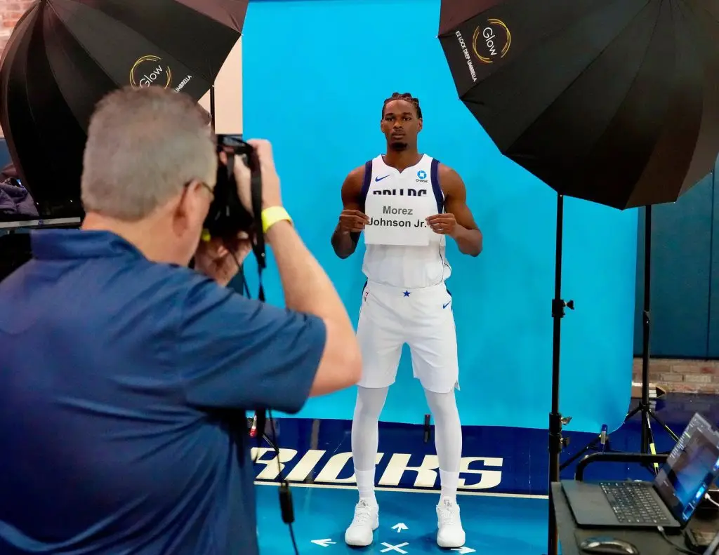 Photographer photographing a Dallas Mavericks player holding a Morez Johnson Jr. identification card in front of a blue backdrop with professional lighting