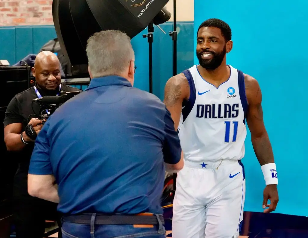 Dallas player in a white number 11 jersey greeting the photographer at the headshot set while a videographer films nearby