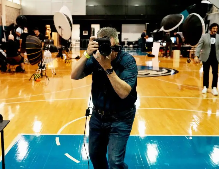 Photographer with camera on an indoor basketball court with professional headshot lighting setups and media crews in the background
