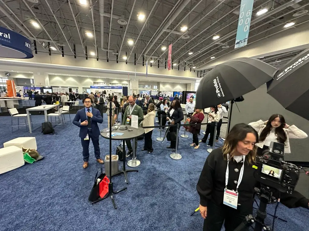 Wide view of a conference headshot station in an exhibit hall: a photographer stands at a tripod-mounted camera in the foreground, an attendee poses between two black umbrella lights, and a line of attendees waits behind stanchions.
