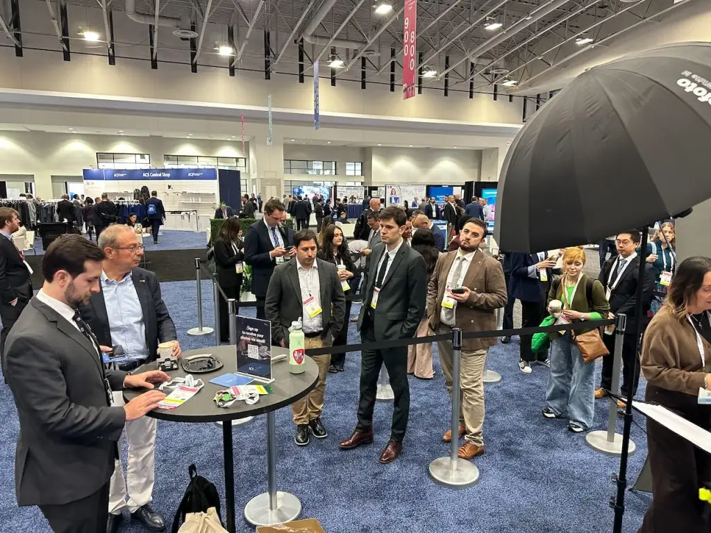 Attendees in business attire gather at a high-top sign-up table and wait in a stanchioned line for headshots, with a black umbrella light visible at right.