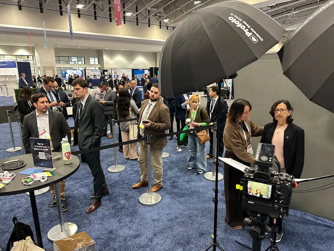 Conference attendees line up between stanchions for headshots in a large exhibit hall while a participant stands at the backdrop in front of a tripod-mounted camera.