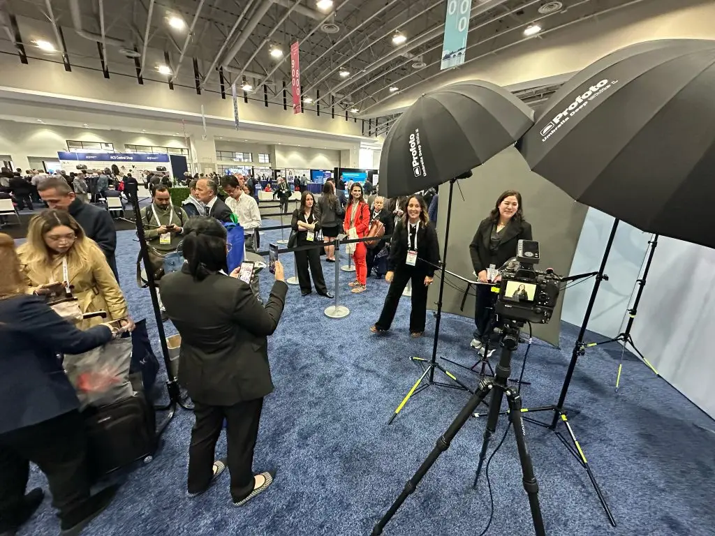 Attendees in a busy exhibit hall wait in line for headshots, some looking at their phones, while a woman poses at a gray backdrop beside a tripod-mounted camera and two umbrella lights.