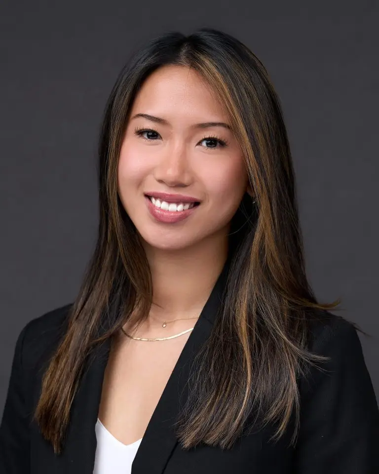 Smiling woman with long dark hair with caramel highlights in a black blazer over a white top and layered gold necklaces.