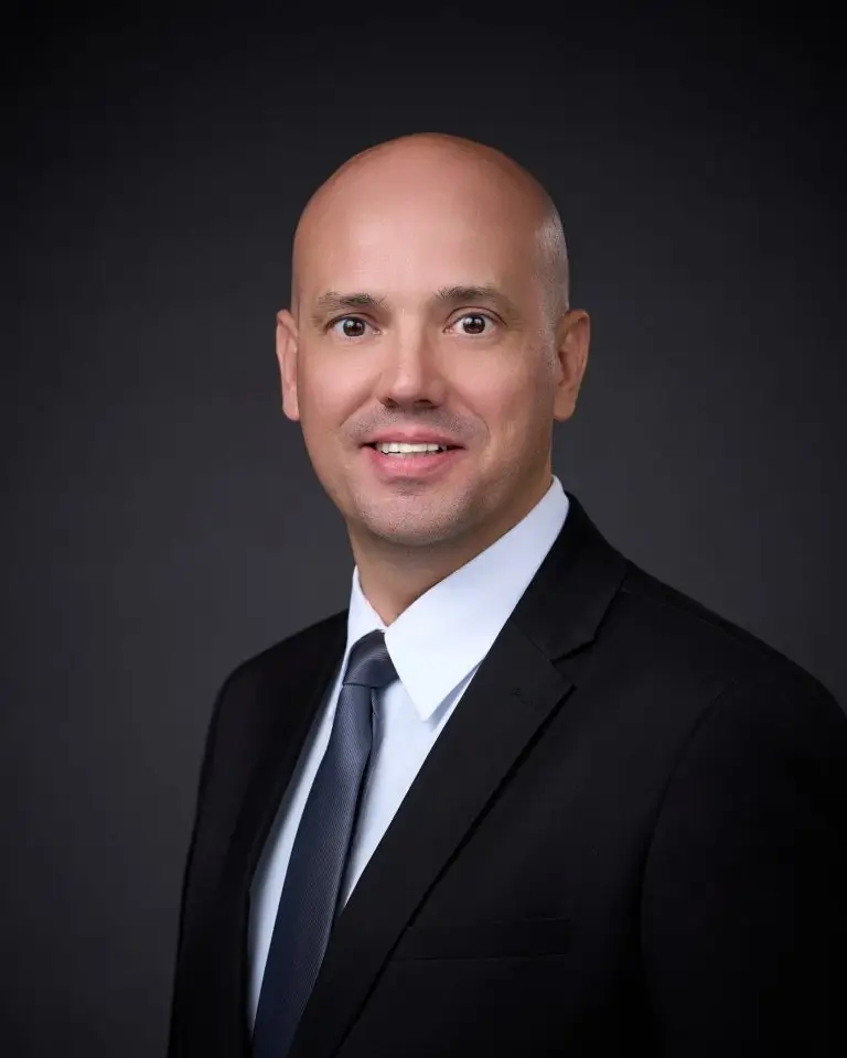 Smiling man with a shaved head wearing a black suit, white shirt and blue-gray tie against a dark gray backdrop.