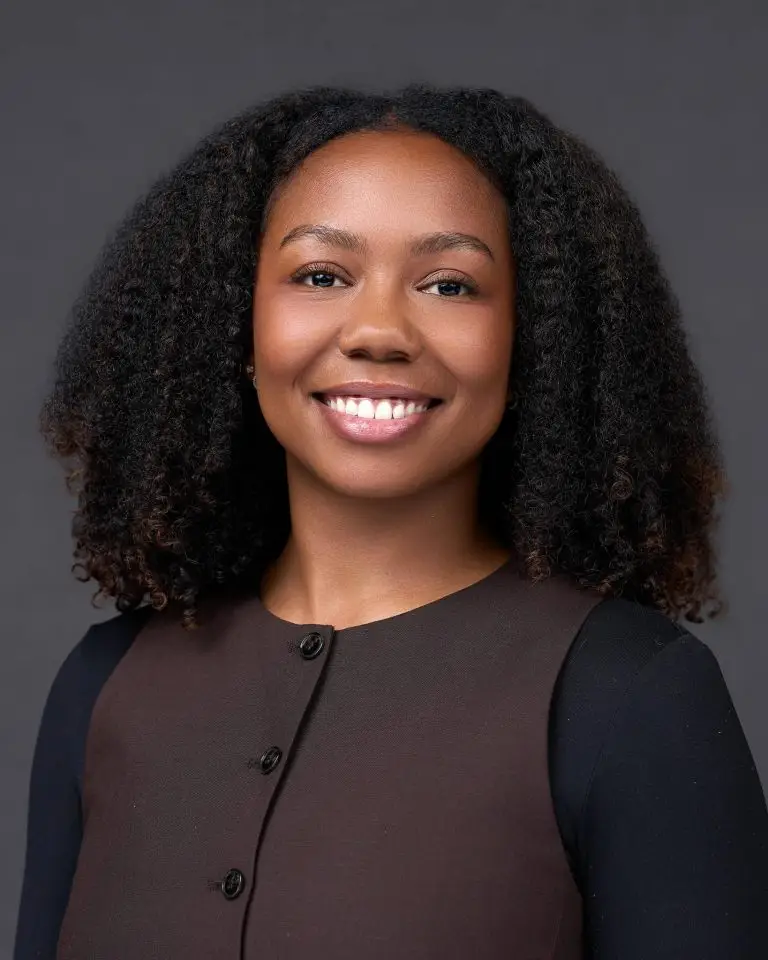 Smiling woman with natural curly hair wearing a brown button-front vest over a black top against a dark gray backdrop.