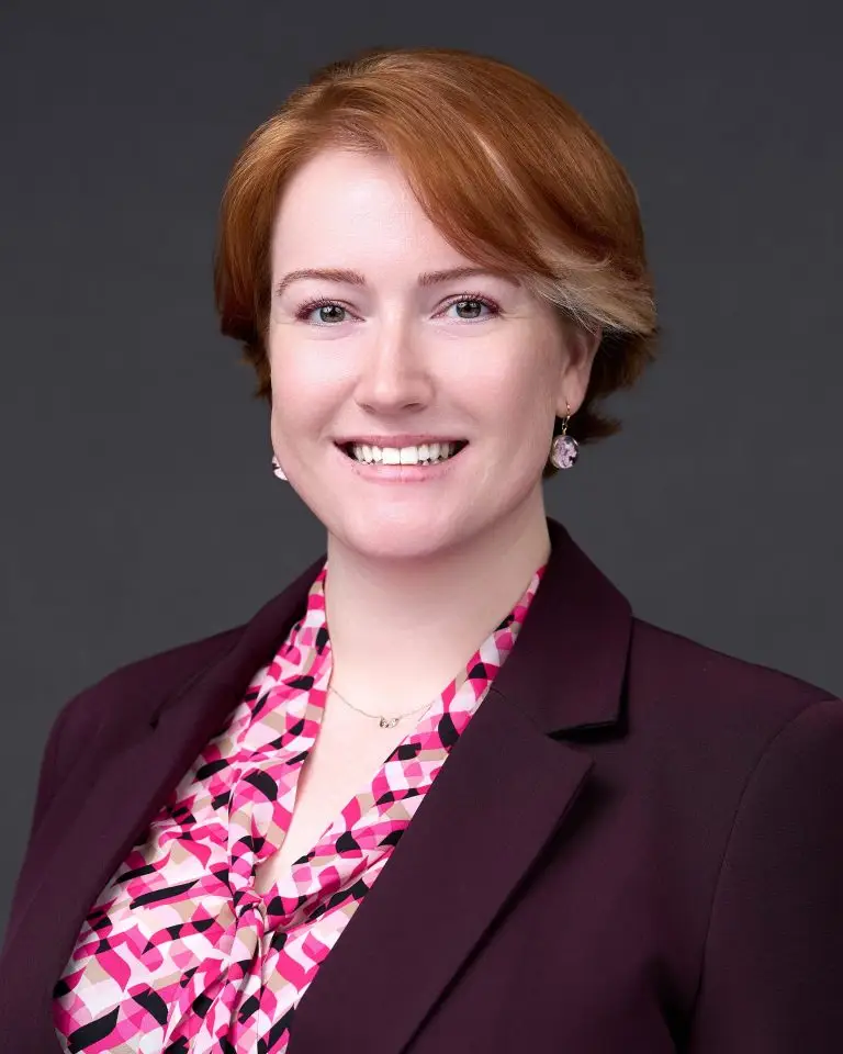 Smiling woman with short auburn hair wearing a burgundy blazer over a pink floral blouse with pearl earrings.