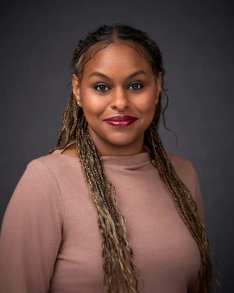 Smiling woman with long highlighted braids wearing a dusty rose mock-neck top against a dark gray backdrop.