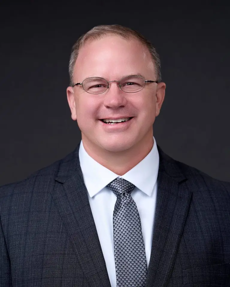 Smiling man with short light hair and glasses wearing a plaid suit, light blue shirt and gray tie.