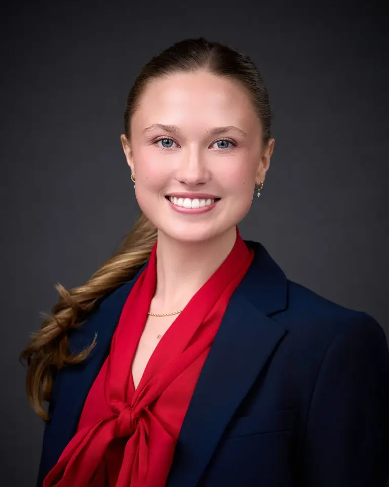 Smiling woman with a long braid over one shoulder wearing a navy blazer and red tie-neck blouse.