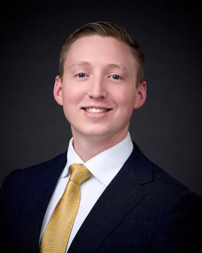 Smiling man with short reddish-blond hair wearing a navy suit, white shirt and gold tie against a dark gray backdrop.