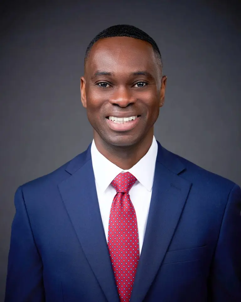 Smiling man with short dark hair wearing a navy suit, white shirt and red tie against a dark gray backdrop.