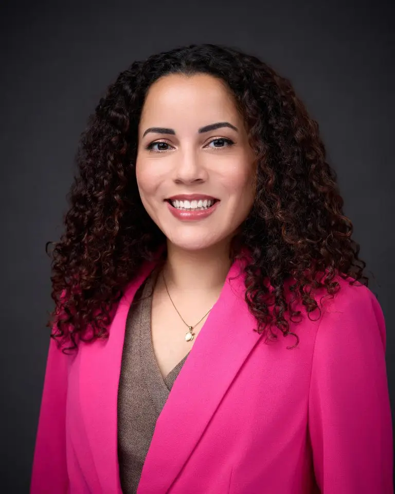 Smiling woman with voluminous curly dark hair wearing a pink blazer over a taupe top against a dark gray backdrop.