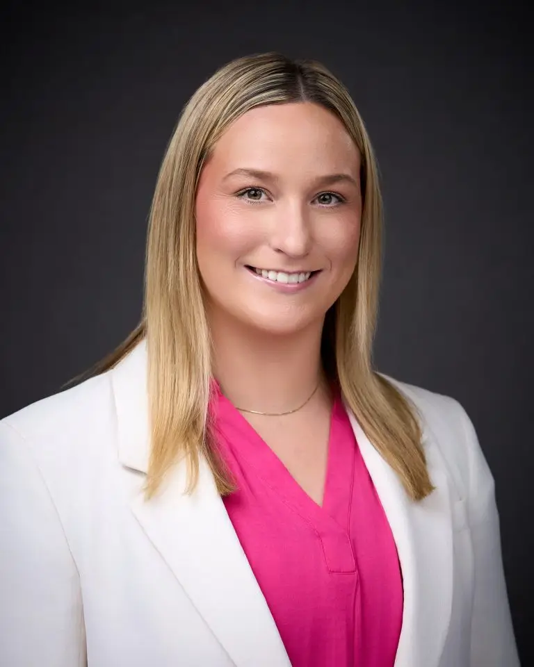 Smiling woman with long blonde hair wearing a white blazer over a pink top with a fine necklace.