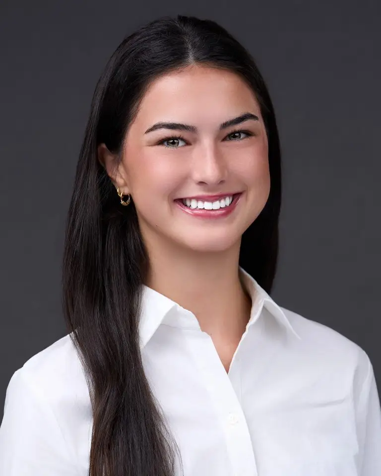 Smiling woman with long dark hair wearing a crisp white collared blouse and gold earrings on a dark backdrop.