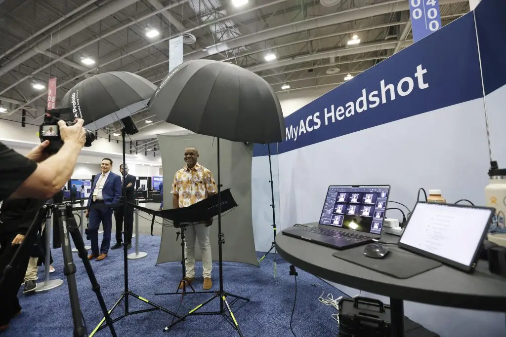 A photographer at left aims a camera at a smiling man in a patterned shirt standing at a gray backdrop between two umbrella lights, as a laptop on a side table shows a grid of headshot previews.