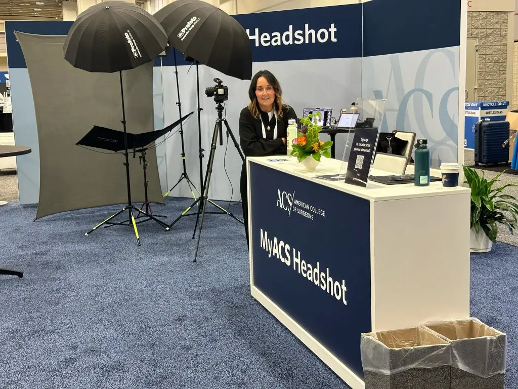 A person sits behind a white MyACS Headshot check-in desk bearing the American College of Surgeons logo, next to a lighting setup with two umbrella lights, a gray backdrop and a camera on a tripod.