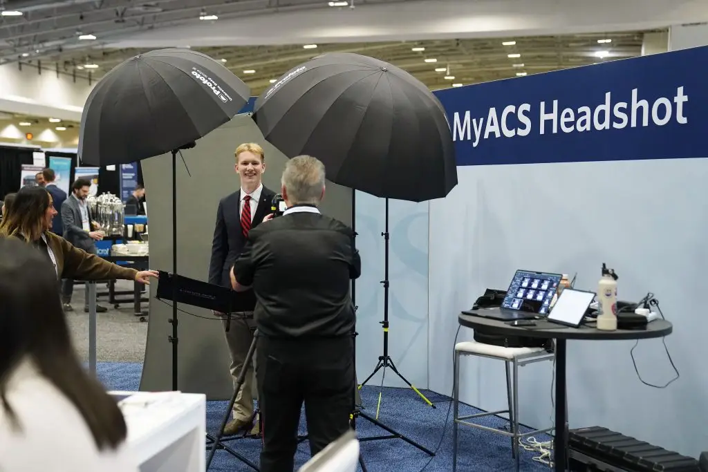 A photographer, seen from behind, photographs a smiling young man in a suit and red tie at a gray backdrop under the MyACS Headshot sign, while a woman holds a reflector at left.
