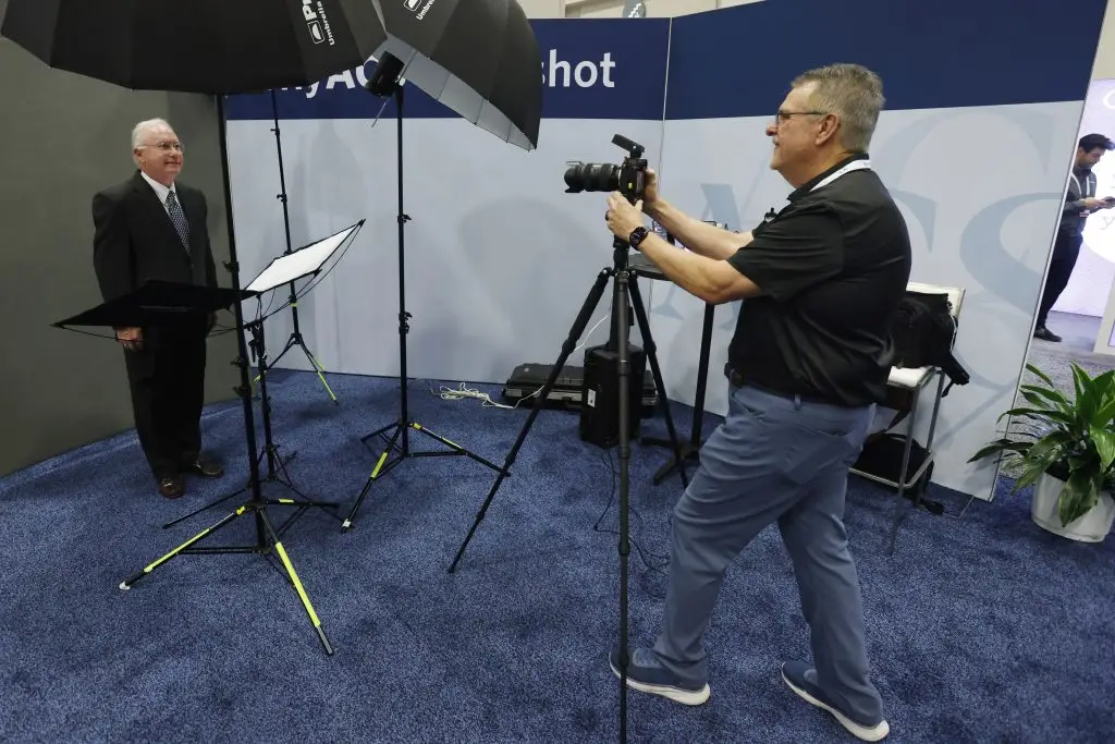 A photographer in a black polo shirt works at a tripod-mounted camera while photographing a man in a dark suit and tie posed between umbrella lights and reflectors.