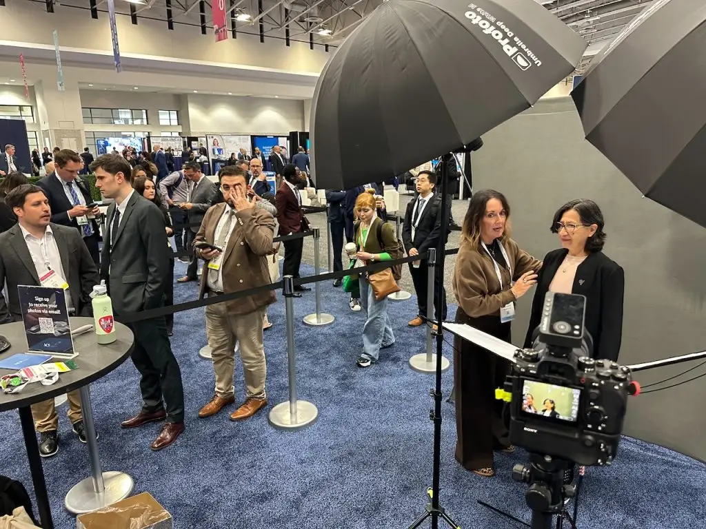 A woman in a tan cardigan helps adjust a participant in a black jacket in front of a gray backdrop while the camera screen shows a live preview and attendees in business attire wait behind stanchions.