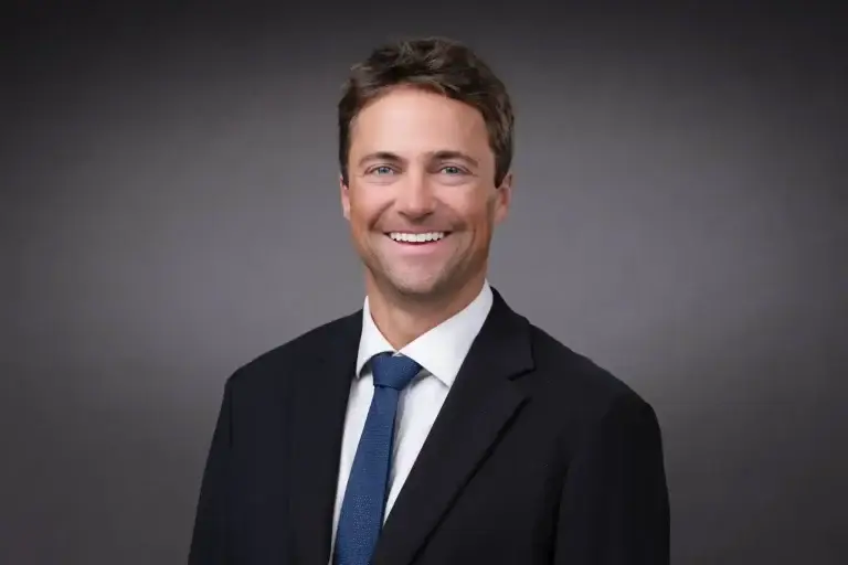 Professional corporate headshot of a smiling business executive wearing a dark suit and blue tie against a gray studio background.